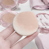 Makeup sponge held by a hand with pink makeup products in the background