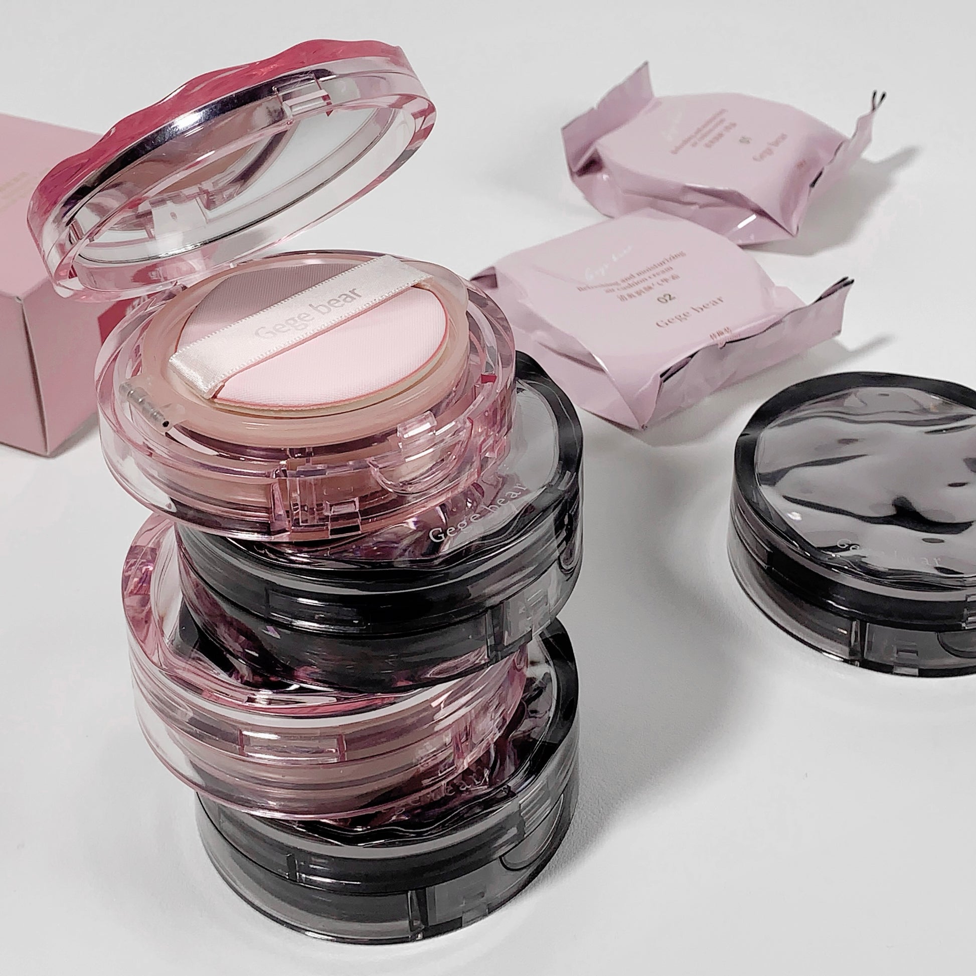 Compact powder in a clear case with pink lid, surrounded by pink packaging on a light background