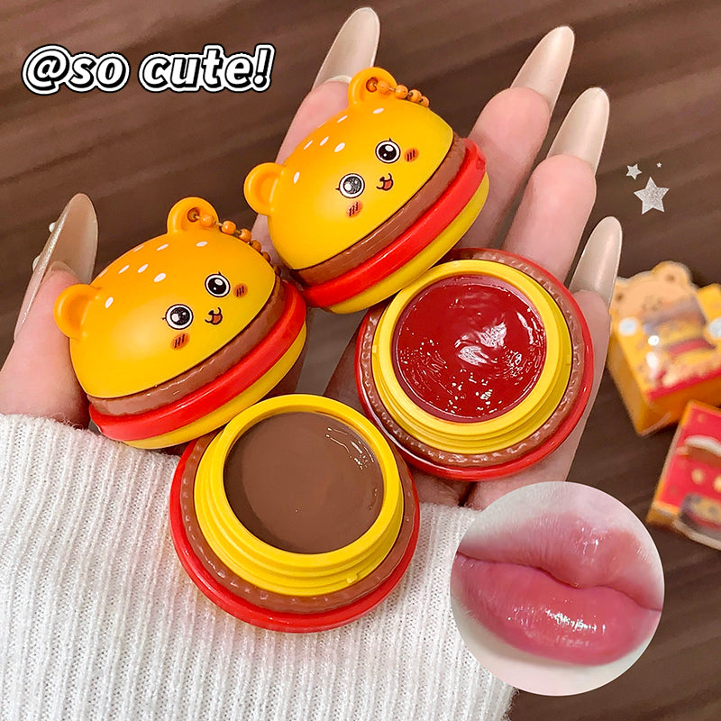 Cute cartoon-style lip balm with a burger design held by a hand, with a close-up of the lip balm and text 'so cute!' on a brown background.