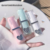 Three lip glosses with heart designs held by a hand on a light surface.
