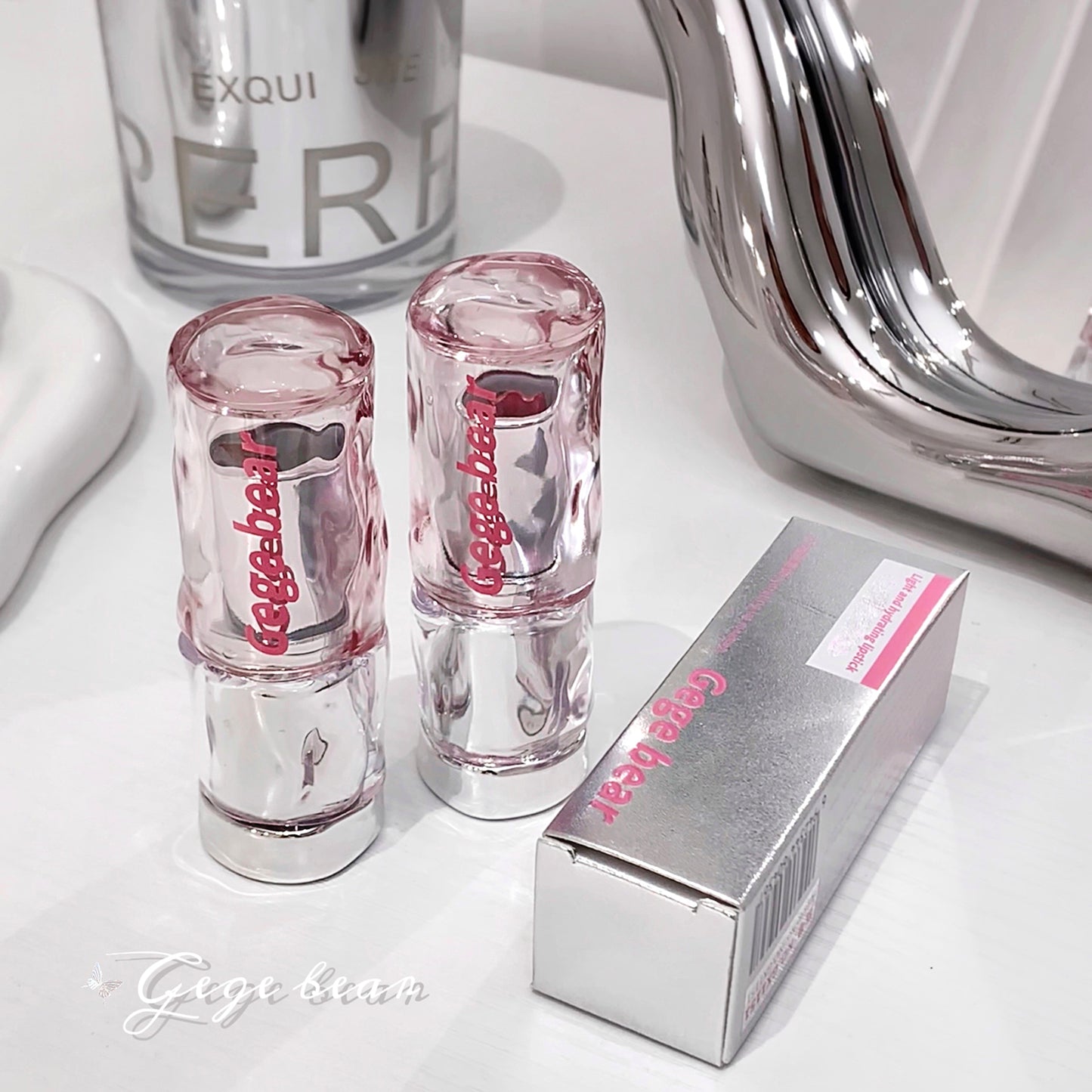 Two pink lip glosses and their packaging on a white surface with a mirror in the background.