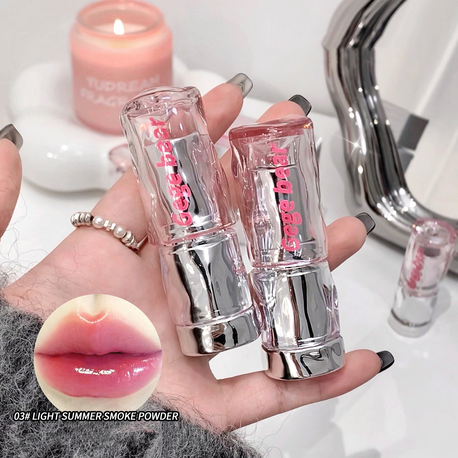 Two lip glosses held in a hand with a candle and mirror in the background