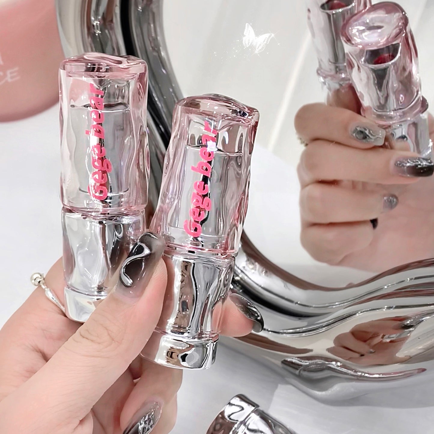 Two clear lip glosses with pink text held by a hand in front of a mirror.