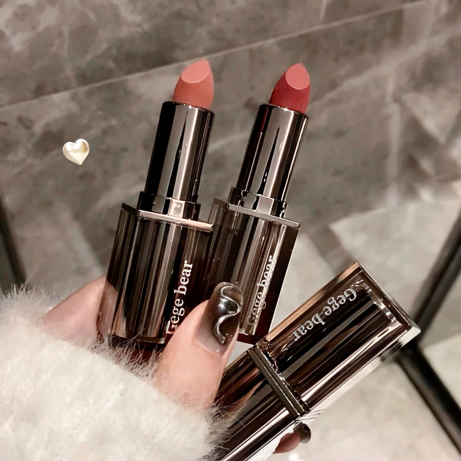 Three lipsticks from the brand 'Age Bear' held by a hand against a neutral background.