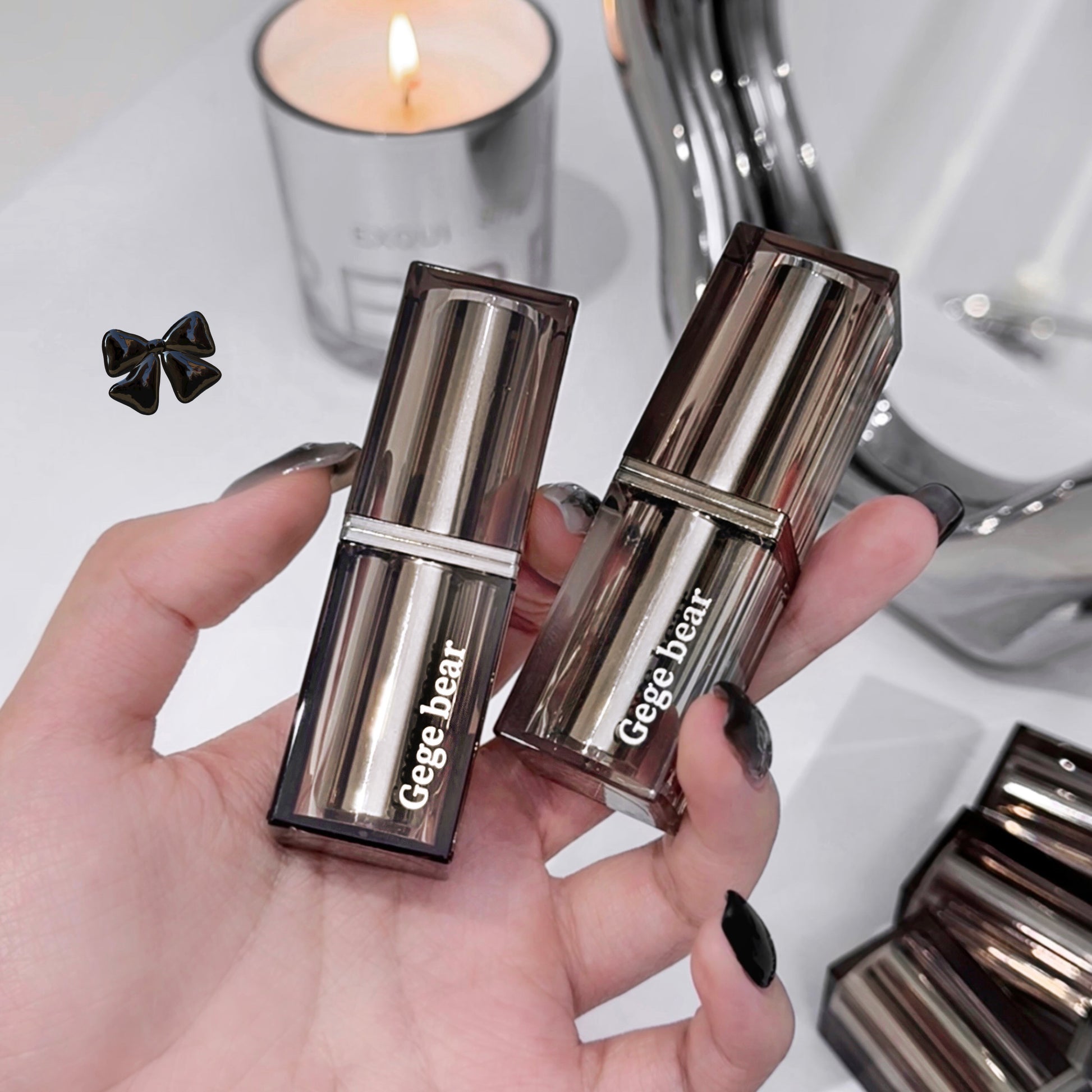 Two metallic Gege bear lipsticks held in a hand with a candle and mirror in the background.