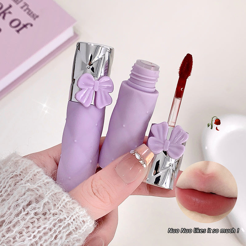 Purple lip gloss with a bow design held by a hand, with a close-up of lips at the bottom right corner.