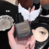 Hand holding a black makeup bottle with angel wings, surrounded by cosmetic items on a dark surface.