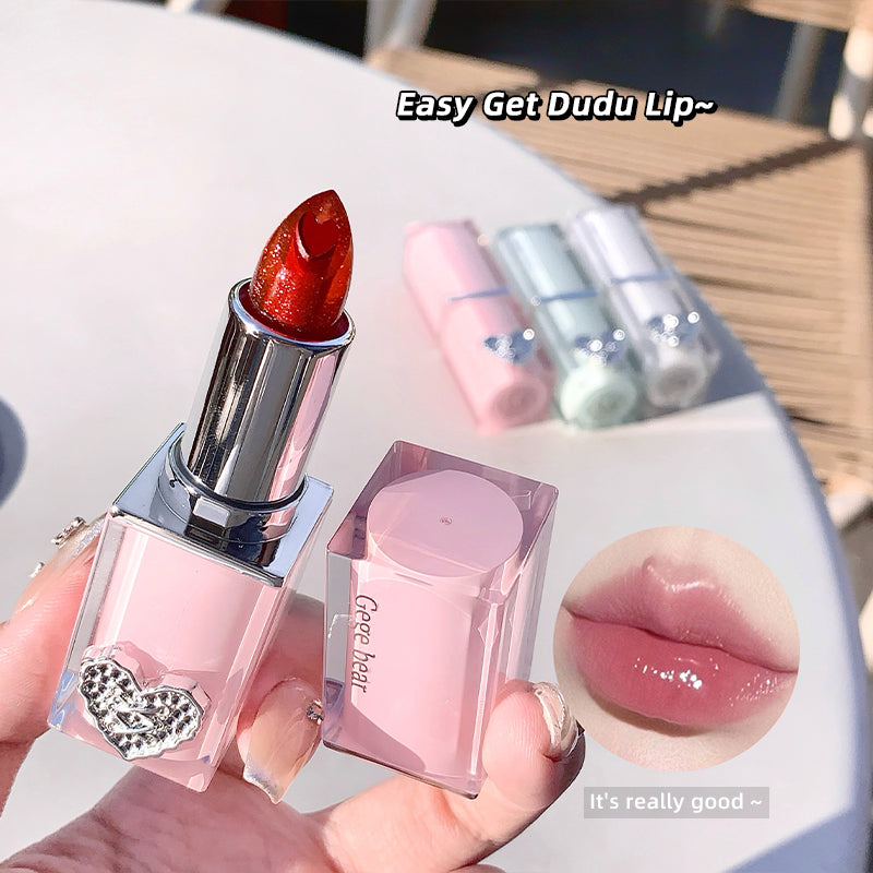 Pink lipstick with a heart detail held by a hand, with a blurred background of more lipsticks.