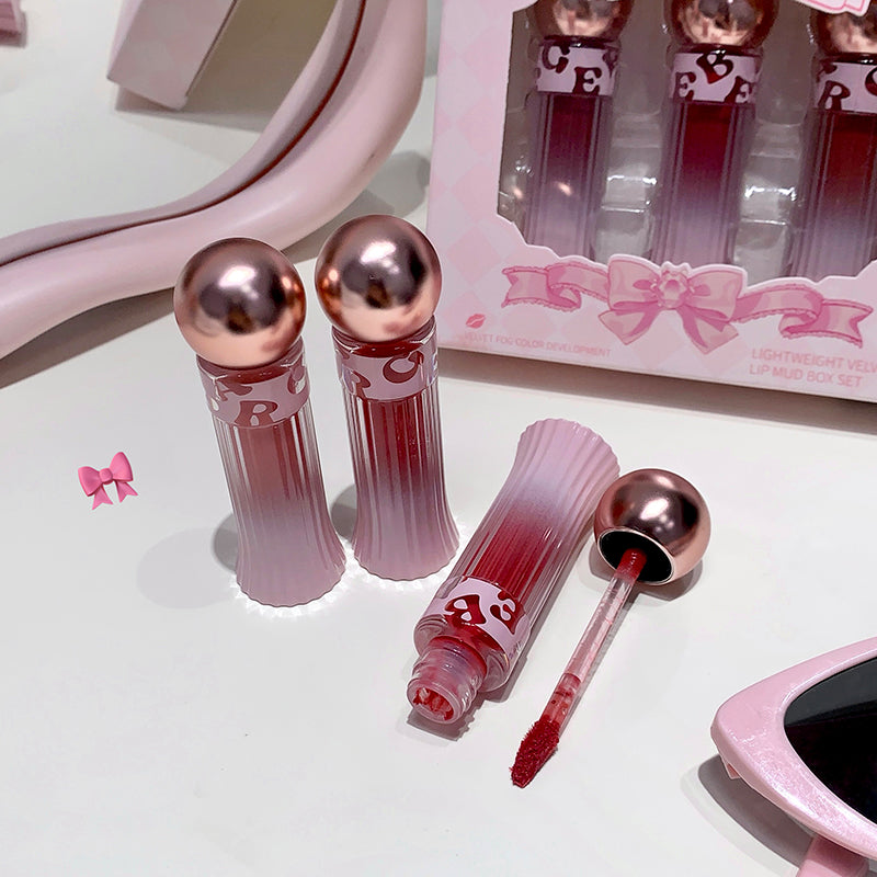 Pink lip gloss with rose gold caps on a white surface, with packaging in the background.