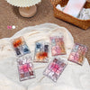 Five small packages of lip gloss on a textured fabric surface with a basket and decorative items in the background.