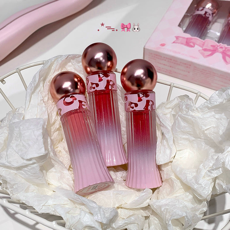 Three pink and red gradient lip glosses with rose gold caps on a white floral background.
