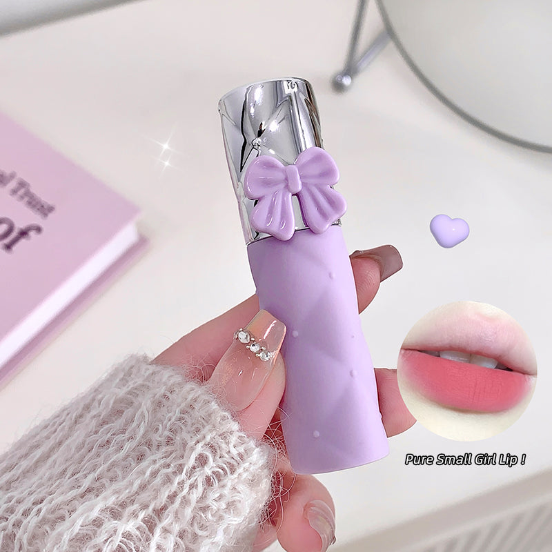 Purple lipstick with a bow design held by a hand, with a close-up of the lipstick color on a pink background.