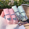 Three lip glosses in pink and green packaging with decorative hearts held by a hand.