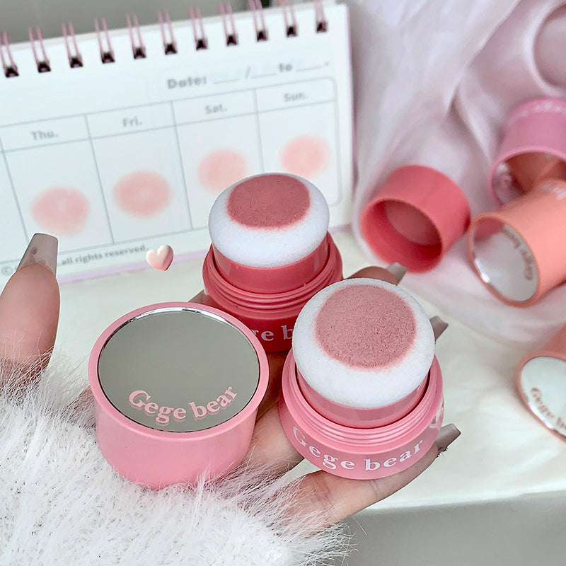Pink makeup sponges with 'Gege bear' branding held by a hand, with a blurred calendar in the background.