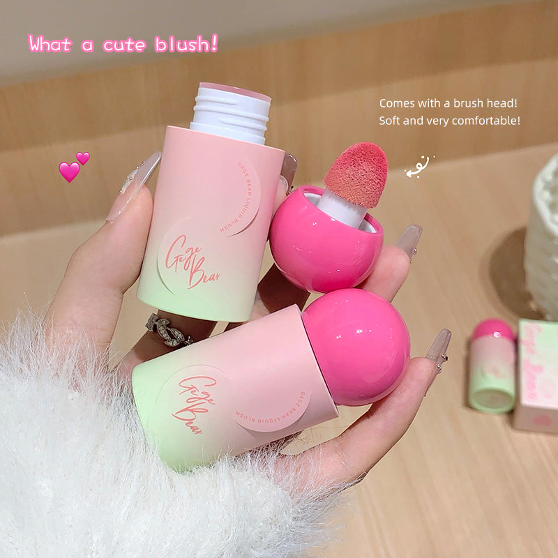 Two pink blushes with a brush head held by a hand on a wooden surface.
