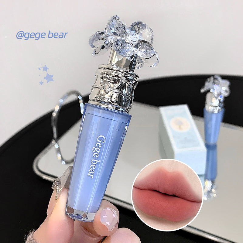 Blue lip gloss with decorative top held by a hand, close-up of lips shown, on a reflective surface.