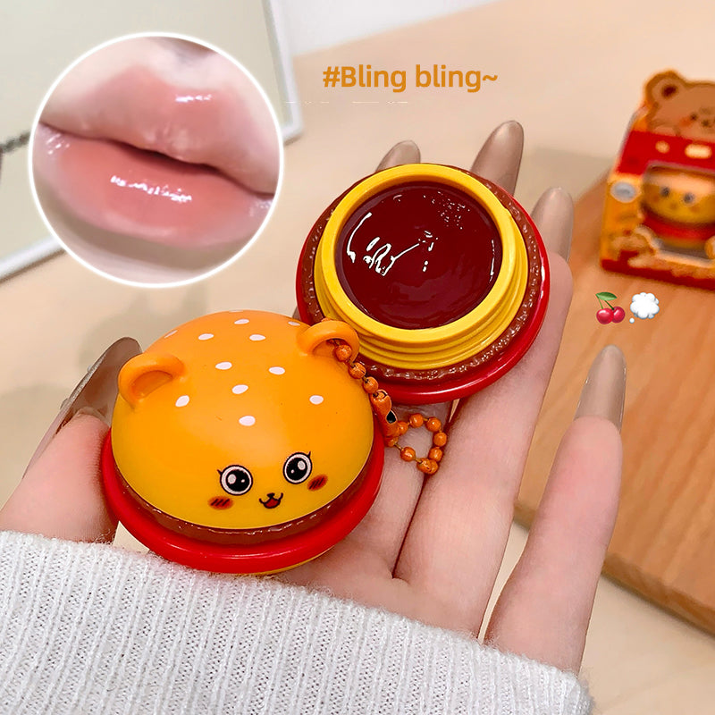 Hand holding a small round container with a cartoon design, showing glossy lip balm inside.