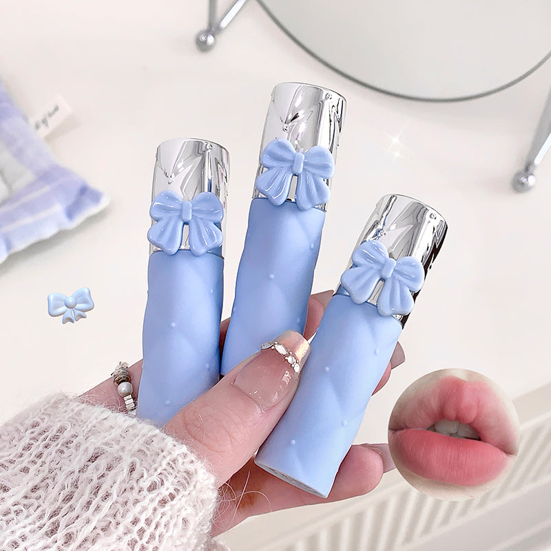 Three blue lip glosses with silver bows held by a hand, with a close-up of lips wearing one of them.