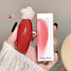 Red lip gloss with white cap held by a hand, next to its packaging, on a neutral background.