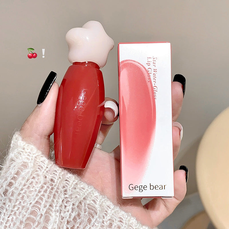 Red lip gloss with white cap held by a hand, next to its packaging, on a neutral background.