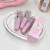 Three pink lip glosses with decorative caps on a white tray, accompanied by their packaging.