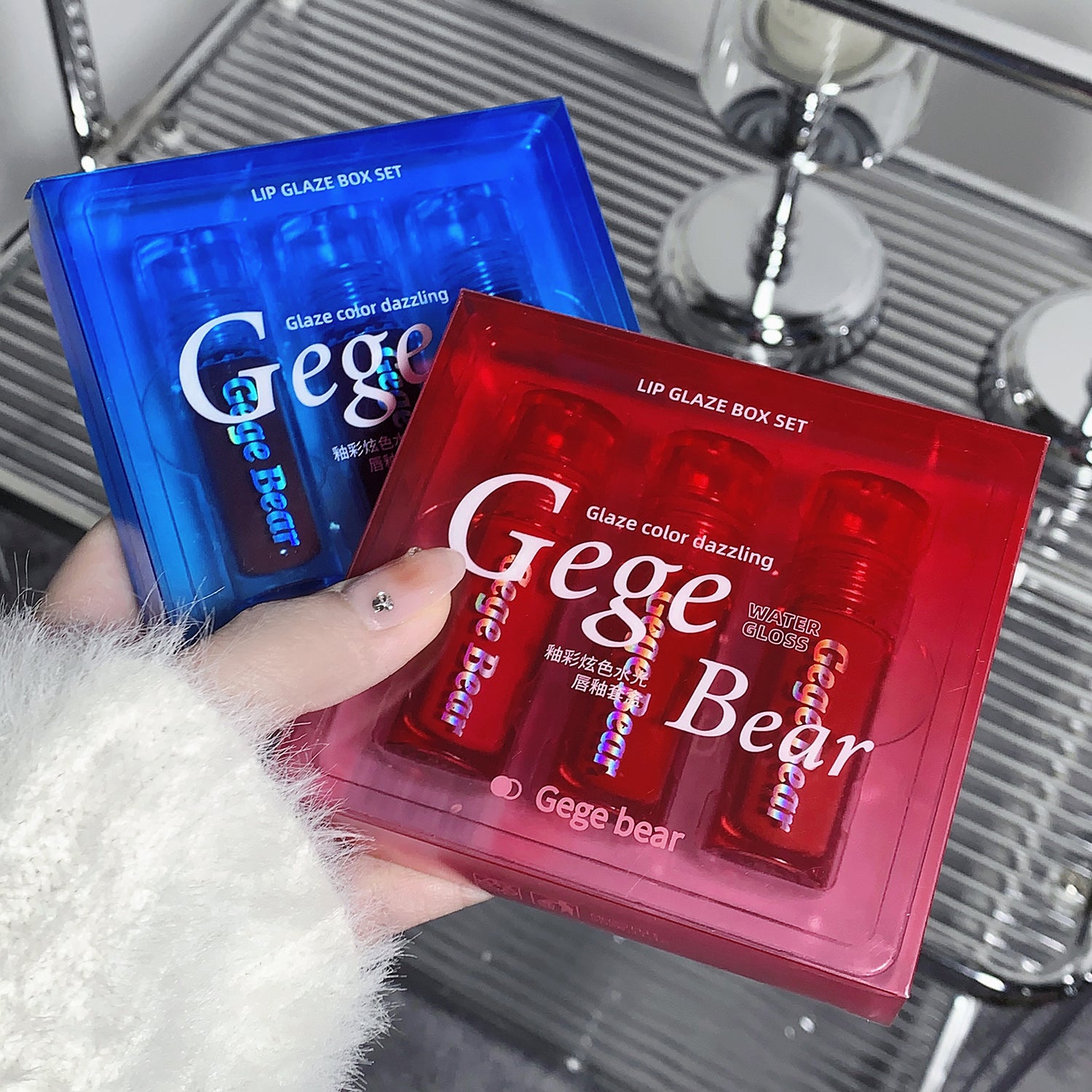 Two lip gloss box sets, one blue and one red, held by a hand with a blurred background.