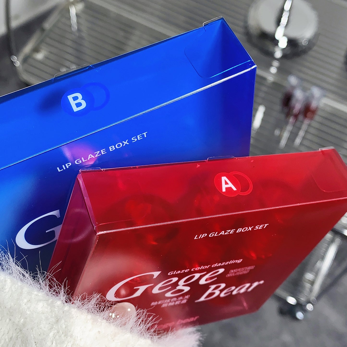 Two lip glaze box sets, one blue and one red, with 'Gege & Bear' branding on a reflective surface.