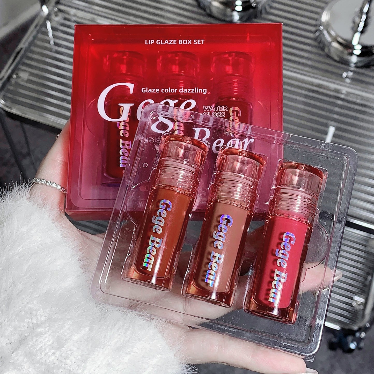Set of lip glosses in clear packaging with 'Gige Bear' branding, held by a person.