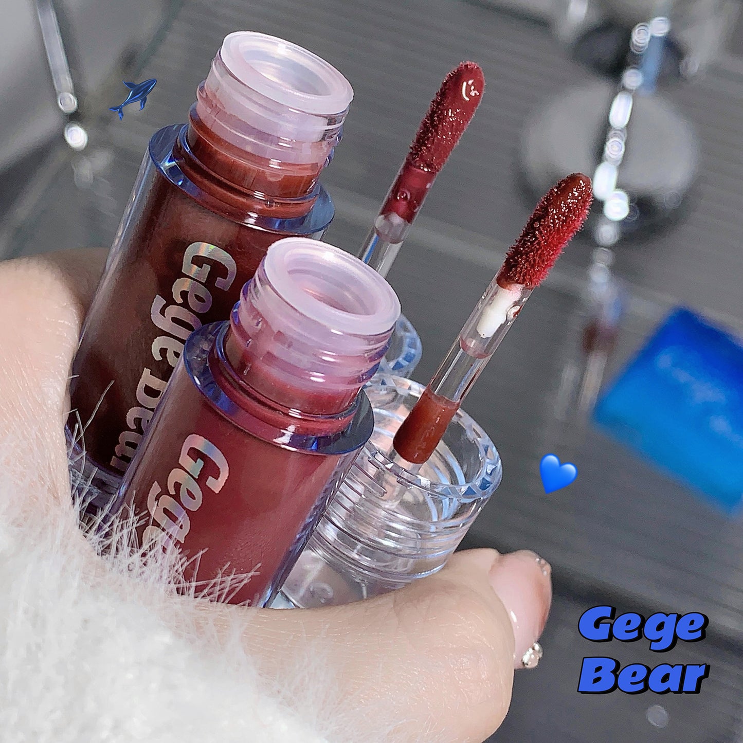 Two lip gloss bottles with applicators held by a hand, blurred bathroom background.