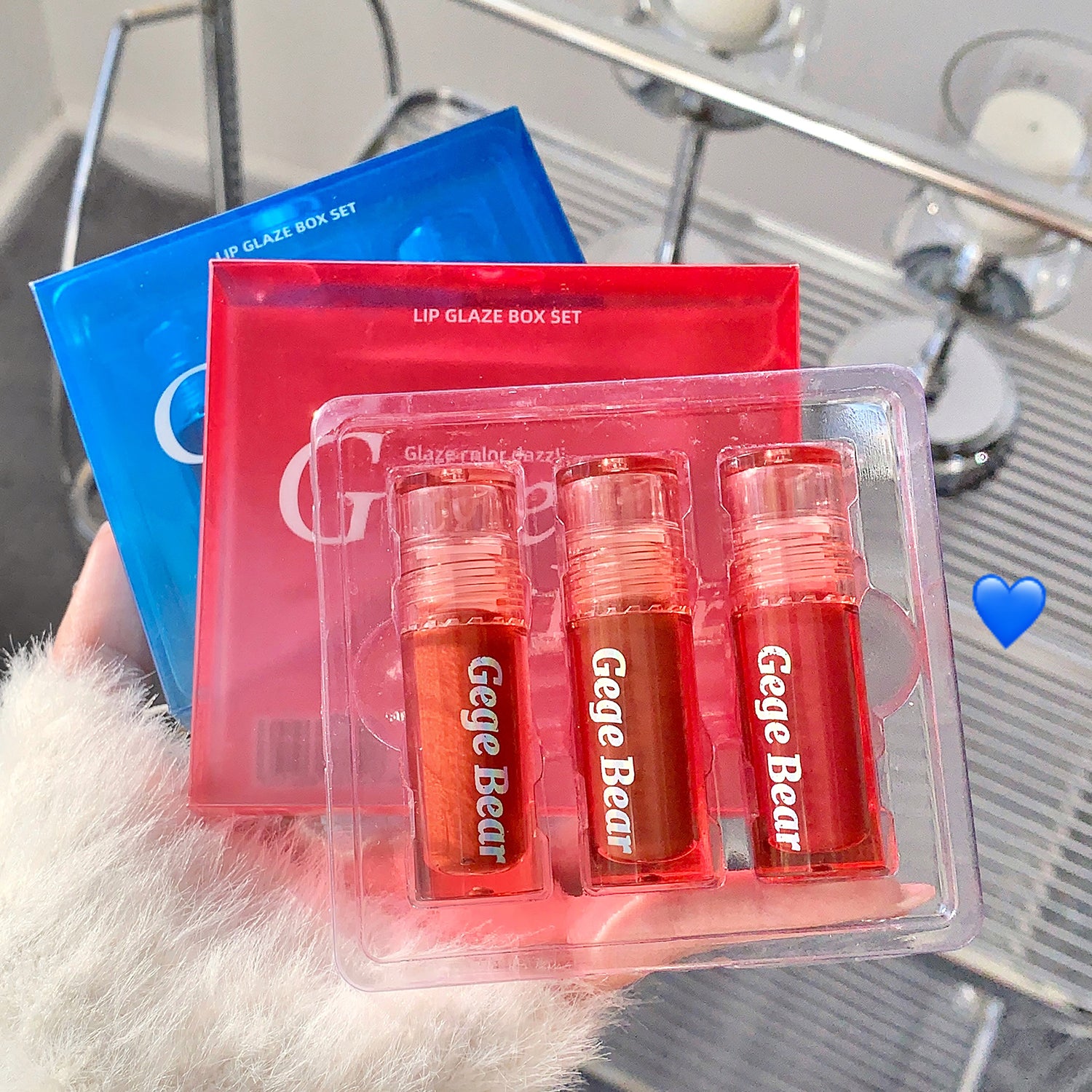 Three lip glosses in a clear packaging with 'Gege Bear' branding, held against a blurred background.