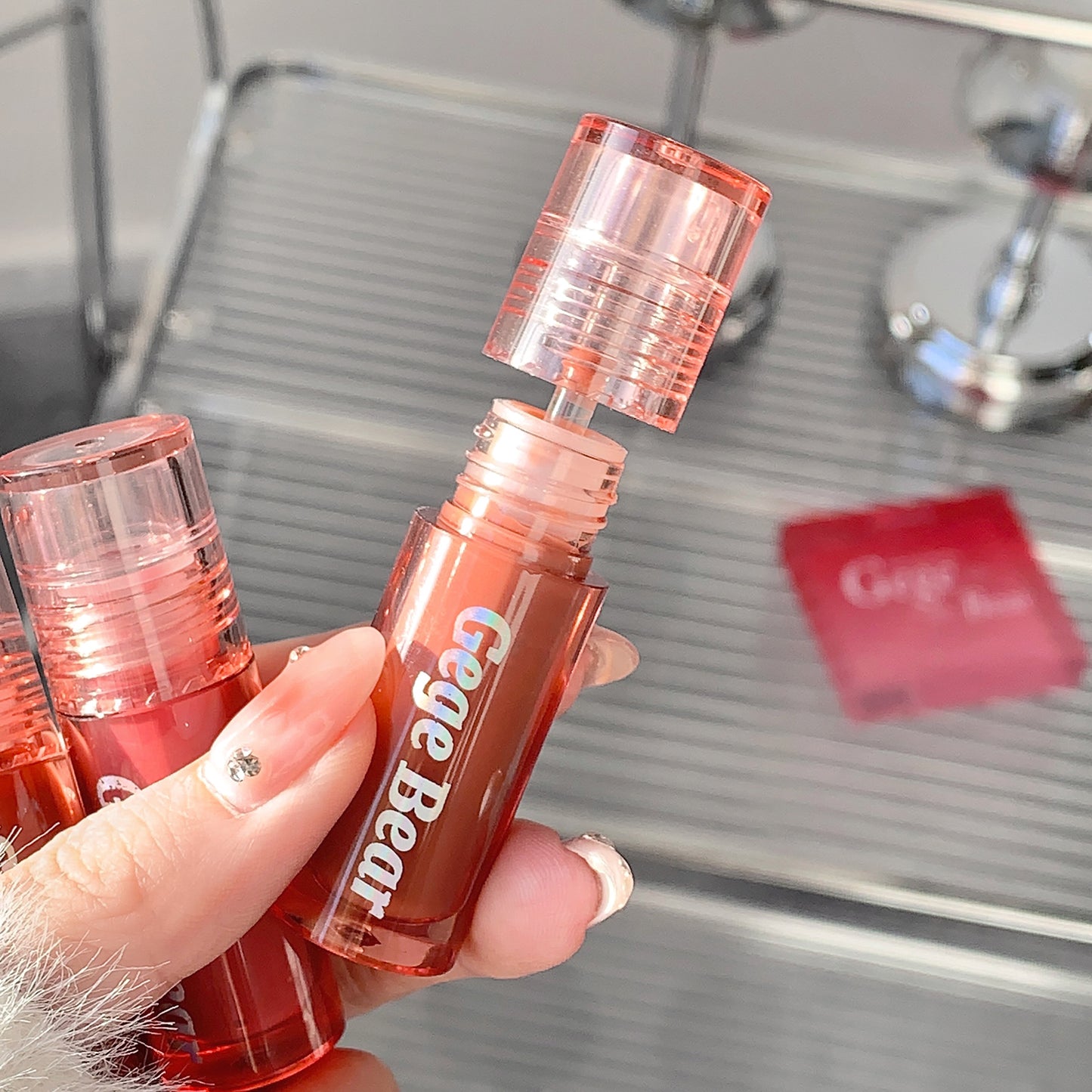 Hand holding a red lip gloss with 'Crege Bear' branding against a blurred background.