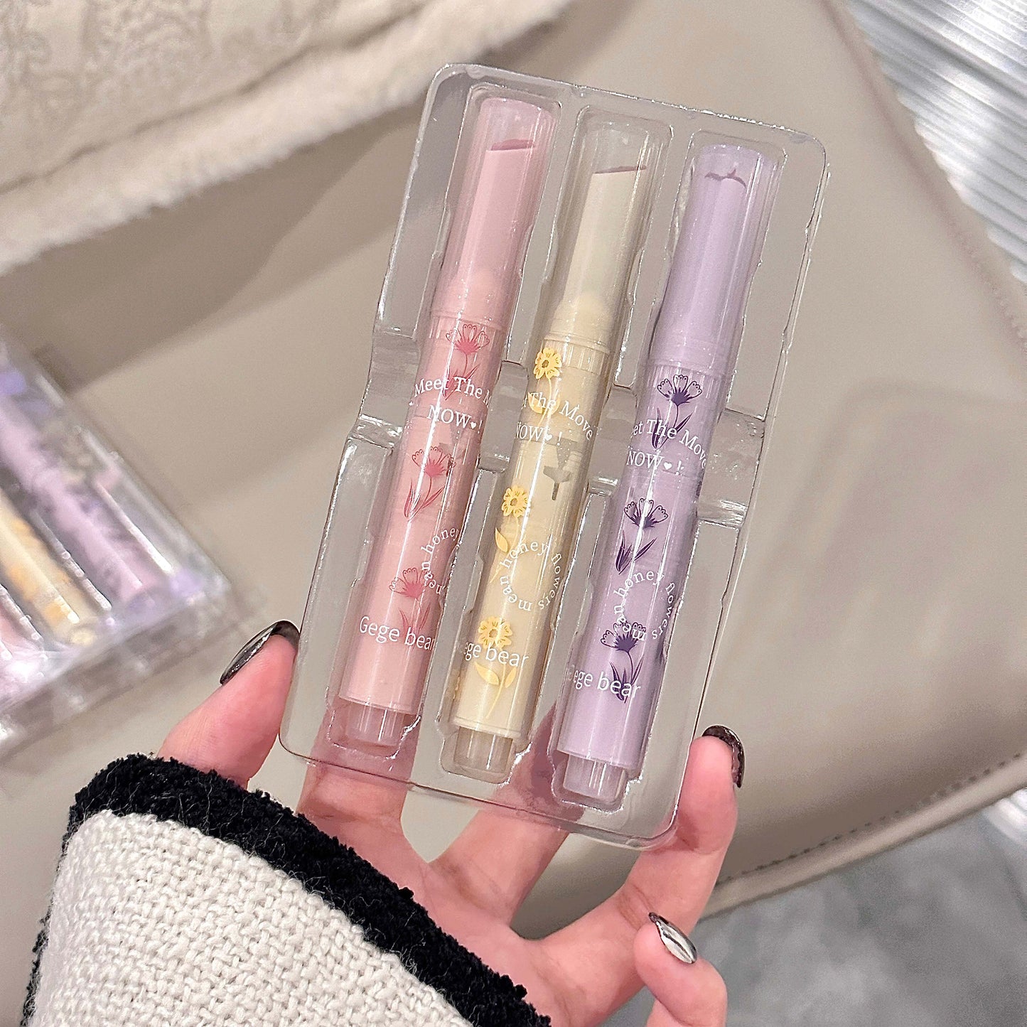 Three lip glosses in clear packaging held by a hand against a neutral background