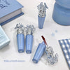 Blue lip gloss with decorative cap on a light surface, surrounded by books and a box.