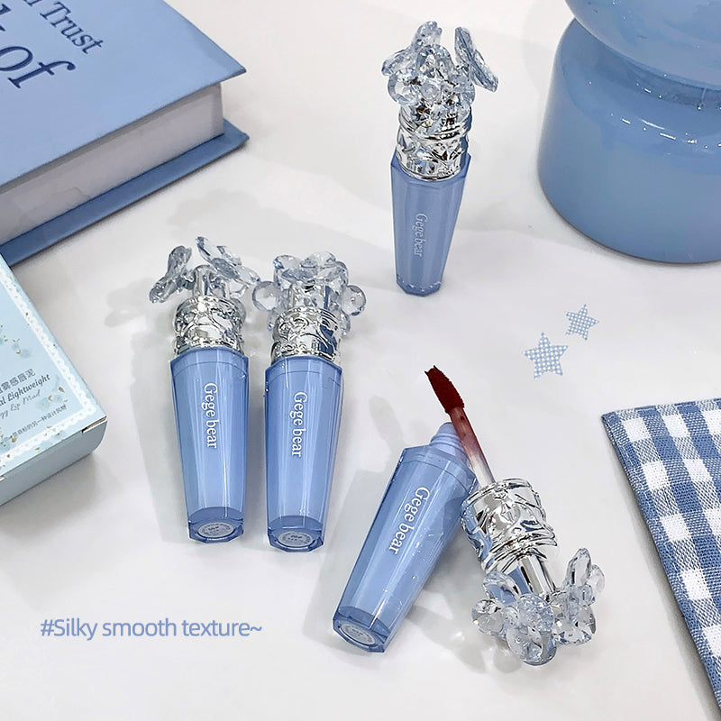 Blue lip gloss with decorative cap on a light surface, surrounded by books and a box.