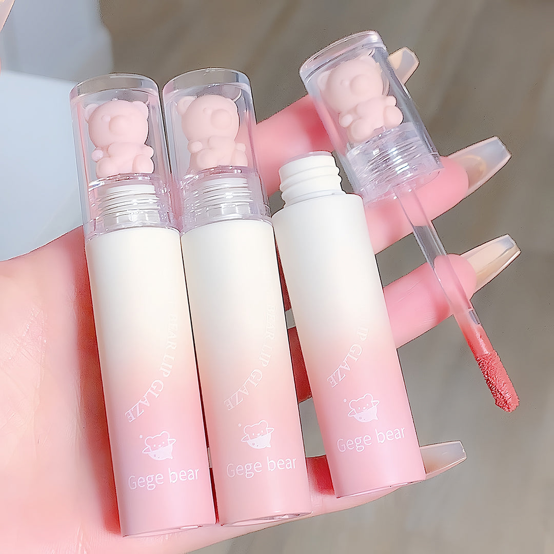 Three lip glosses with bear-shaped caps held by a hand, one with a visible color swatch.