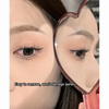 Close-up of a person's eye with false eyelashes, using a heart-shaped mirror to check the application.