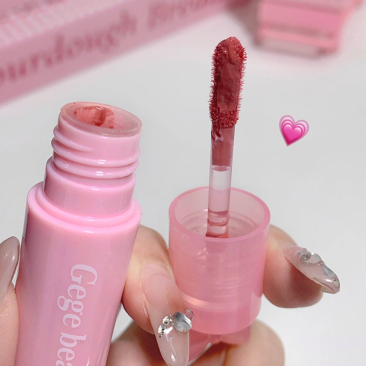 Pink lip gloss with applicator held by a hand, brand 'Geege' visible