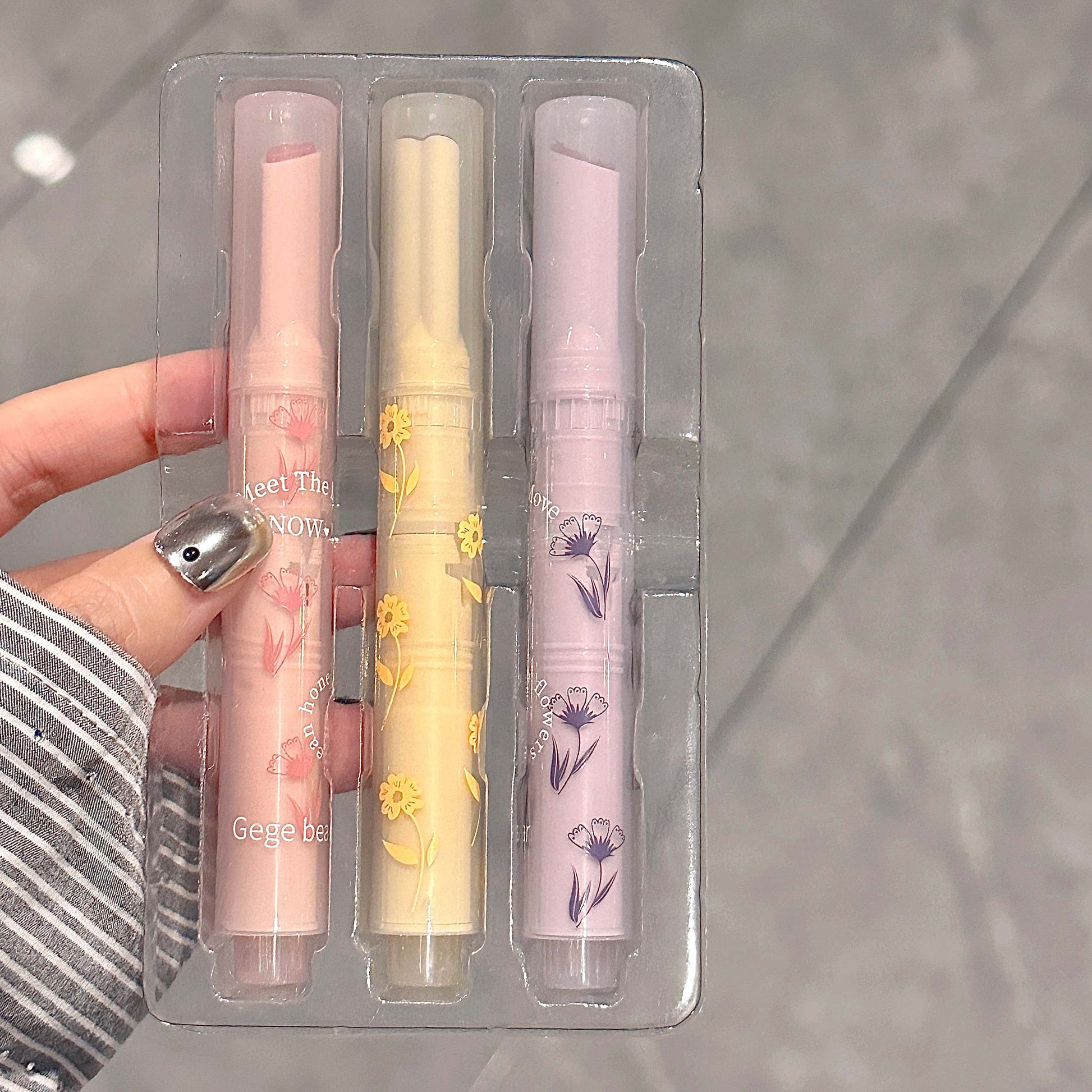 Three lip balms in a clear packaging with floral designs held by a hand.