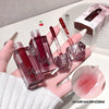 Hand holding four lip glosses with a cherry on top, set against a neutral background.