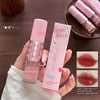 Pink lip gloss with packaging held by a hand on a wooden surface, with additional lip glosses in the background.