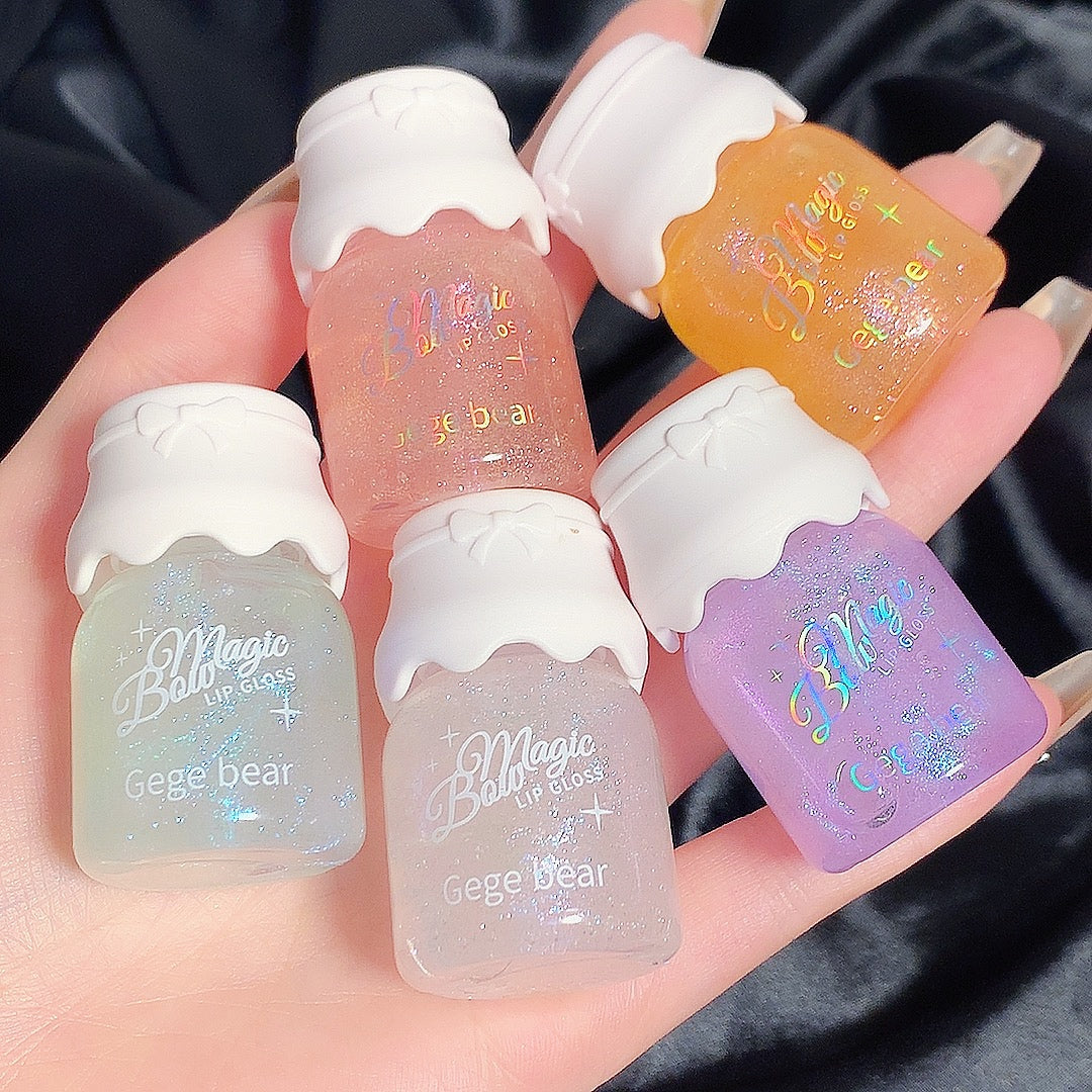 Five small lip gloss bottles with colorful labels held in a hand against a dark background.