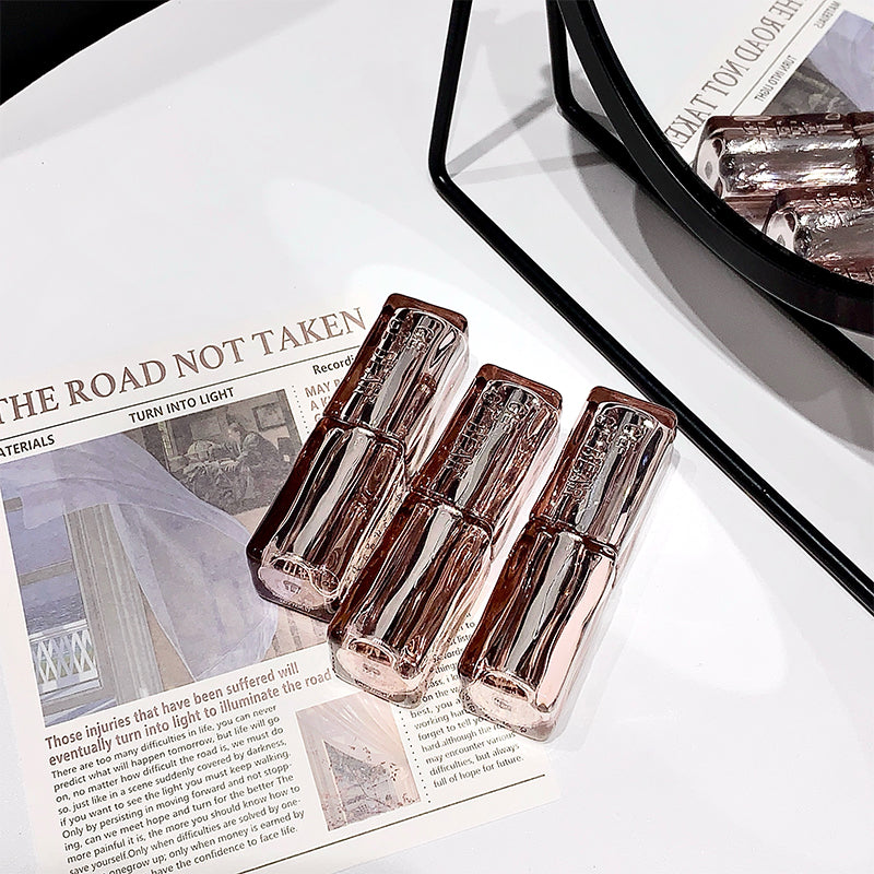 Three transparent lip glosses on a newspaper with a mirror and pink items in the background.