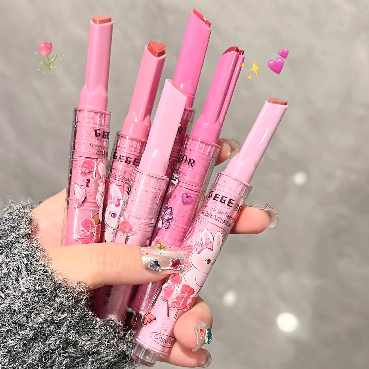 Set of pink lip products with cartoon designs held by a hand against a gray background.