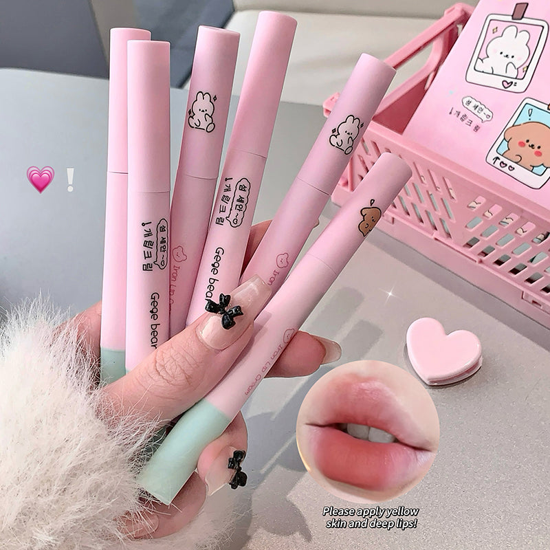 Pink lip glosses with cartoon designs held by a hand, with a pink basket and heart-shaped object in the background.