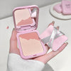 Pink makeup compact with mirror held by a hand, close-up of eye, and blurred background.