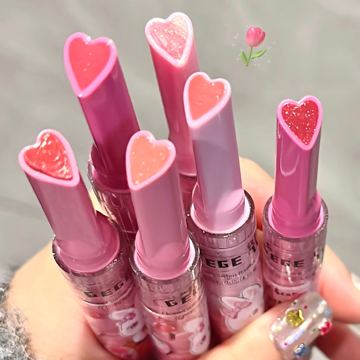 Set of pink lip balms with heart-shaped caps held by a hand.