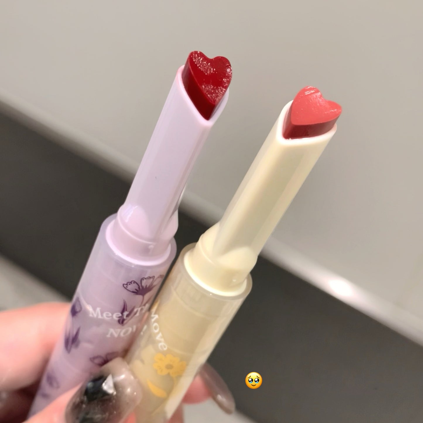 Two lip glosses with heart-shaped caps held by a hand against a neutral background