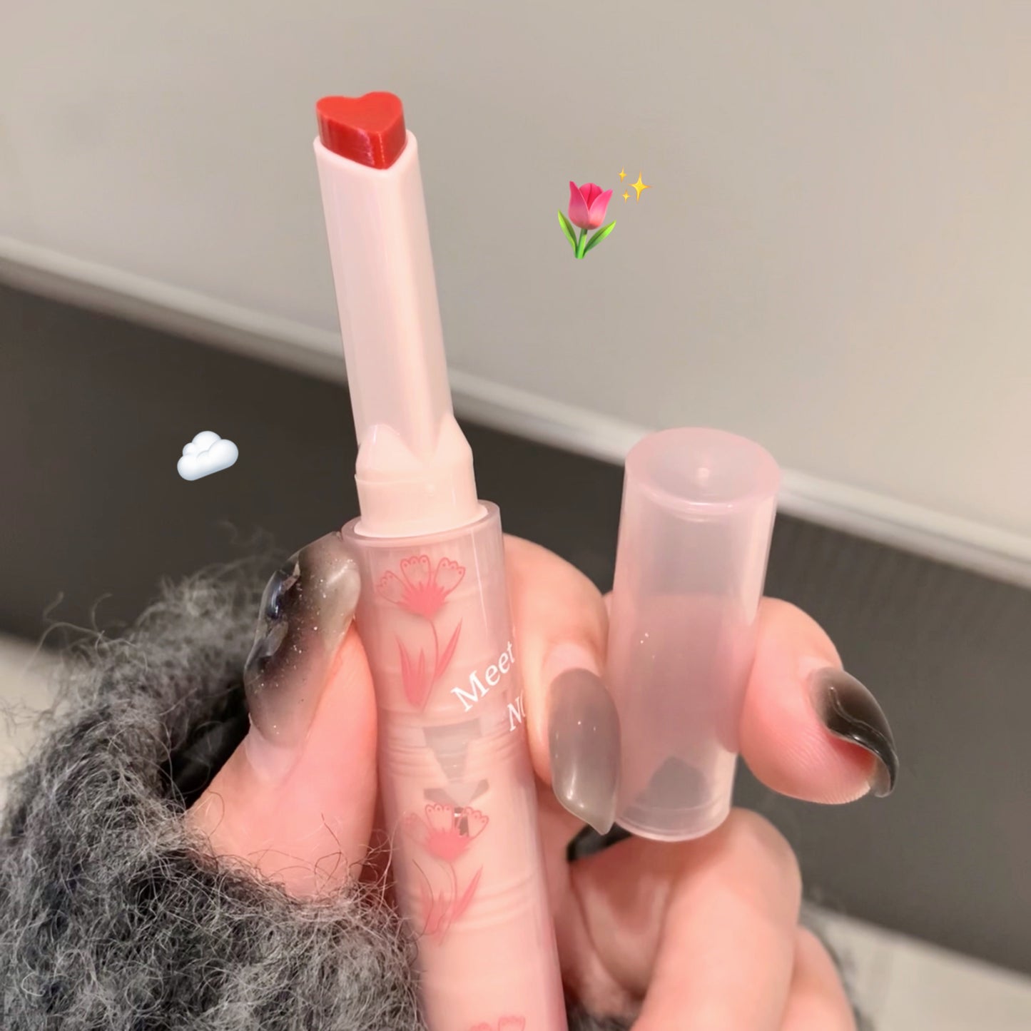 Hand holding a small pink lipstick with a heart-shaped cap against a neutral background