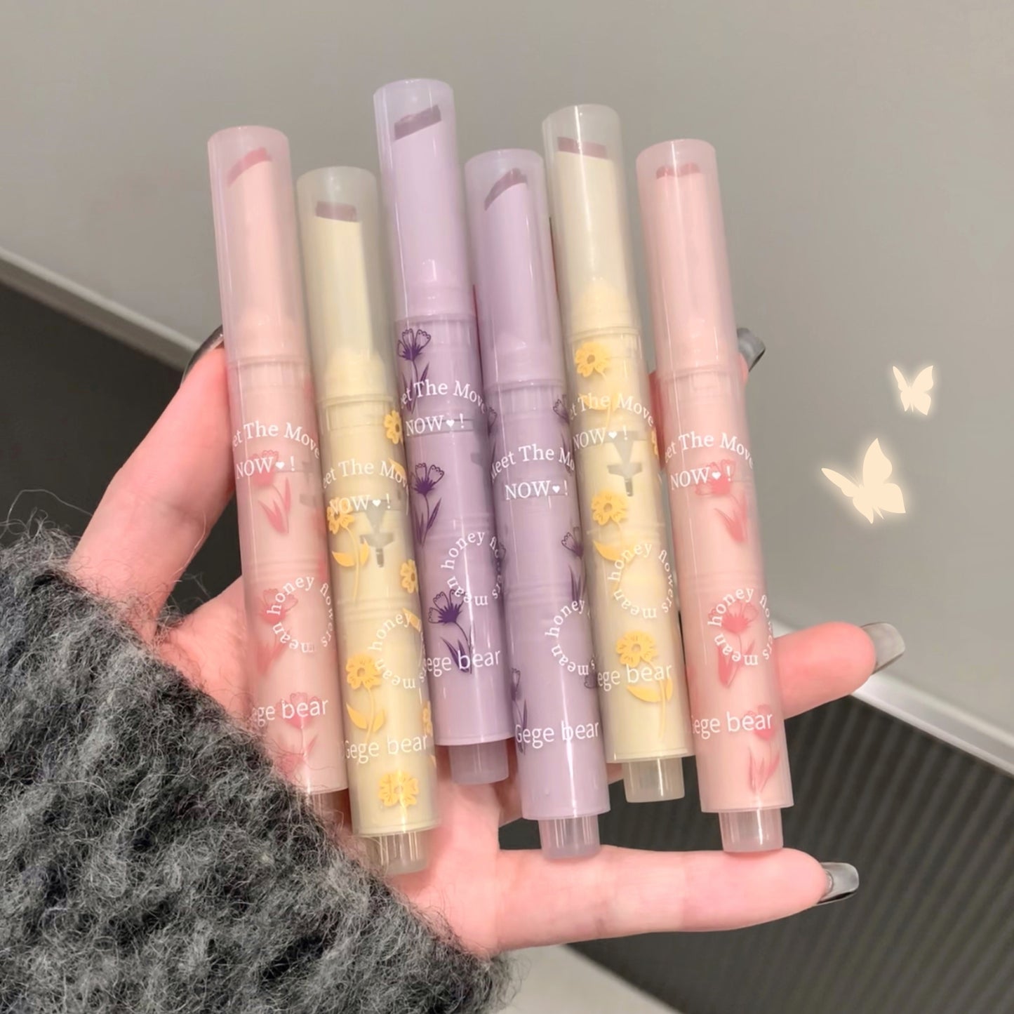 Hand holding six lip balms with floral designs against a neutral background