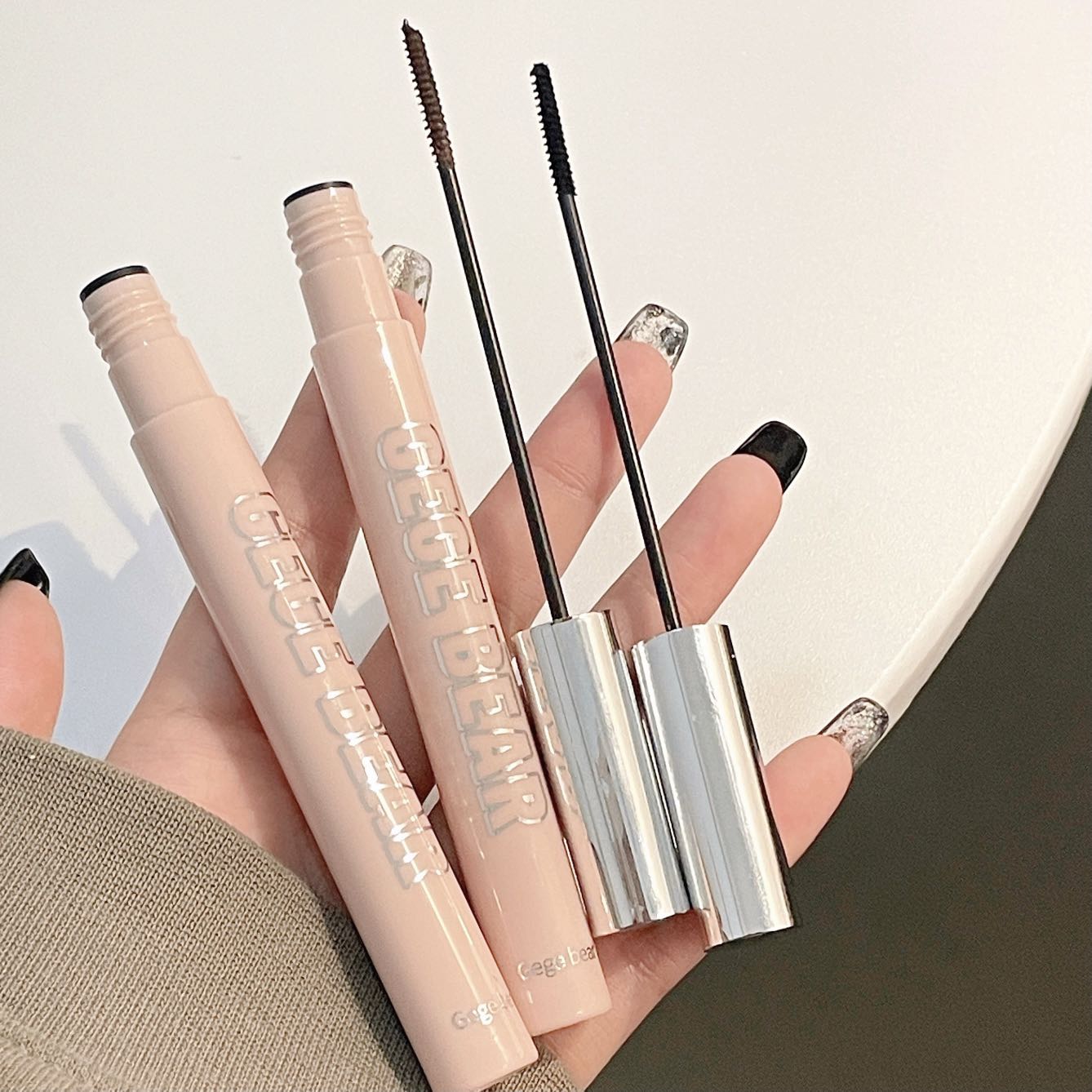 Makeup products including mascara and eyeliner held by a hand on a neutral background